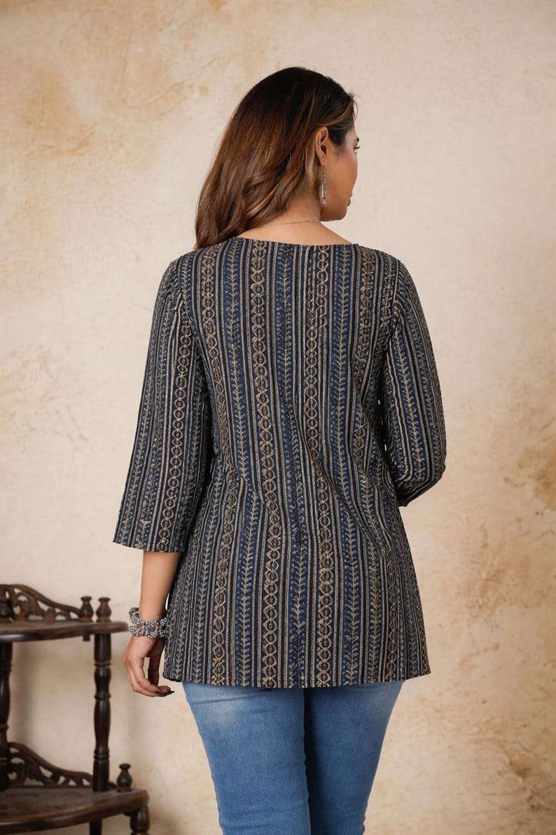 Pu&ograve; includere: Una tunica blu scuro caratterizzata da un motivo verticale di disegni geometrici e a forma di foglia color oro. Il capo presenta maniche a tre quarti e uno scollo rotondo, abbinato a jeans in denim di colore azzurro.