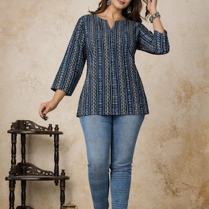 Pu&ograve; includere: Una tunica blu scuro con un motivo a righe verticali bianche e dorate, caratterizzata da maniche a tre quarti e scollo a V, abbinata a jeans azzurri con dettagli di cuciture orizzontali lungo le gambe.
