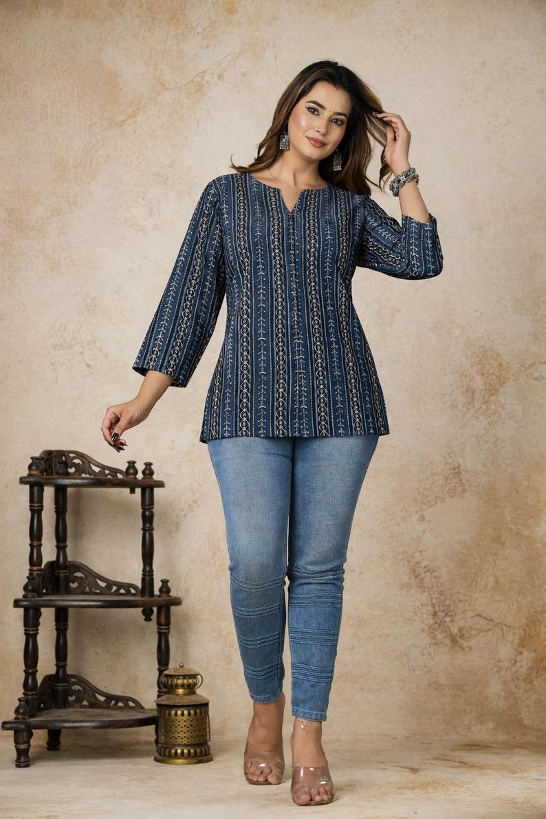 Pu&ograve; includere: Una tunica blu scuro con un motivo a righe verticali bianche e dorate, caratterizzata da maniche a tre quarti e scollo a V, abbinata a jeans azzurri con dettagli di cuciture orizzontali lungo le gambe.