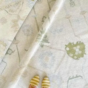 May include: A cream-colored rug with a subtle floral pattern in shades of green and gray. The rug is folded, revealing its texture and design. The image also shows a pair of yellow and white striped shoes.