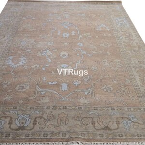 May include: A large, rectangular rug with an intricate floral pattern in shades of tan, light blue, and brown. The rug has a fringe along the bottom edge and the text "VTRugs" is visible on the surface.