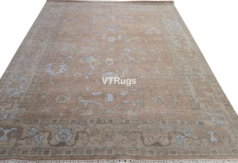 May include: A large, rectangular rug with an intricate floral pattern in shades of tan, light blue, and brown. The rug has a fringe along the bottom edge and the text "VTRugs" is visible on the surface.