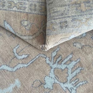 May include: Close-up of a beige rug with a geometric pattern in light blue and gray. The rug is folded over, revealing the underside and the texture of the woven fibers. The design features stylized floral or branch-like motifs.