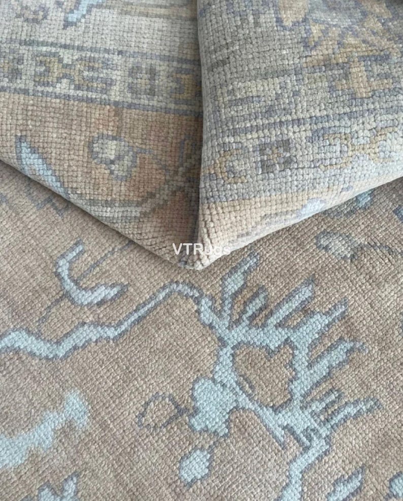 May include: Close-up of a beige rug with a geometric pattern in light blue and gray. The rug is folded over, revealing the underside and the texture of the woven fibers. The design features stylized floral or branch-like motifs.