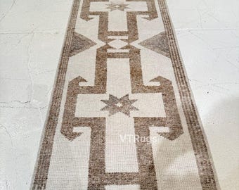 May include: A rectangular runner rug with a cream-colored background and a repeating geometric pattern in shades of brown. The design includes cross-like shapes and star motifs, with a fringed edge.