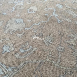 May include: Close-up of a beige rug with a detailed floral pattern in light blue and off-white. The rug has a textured appearance and the text "VTRugs" is visible.