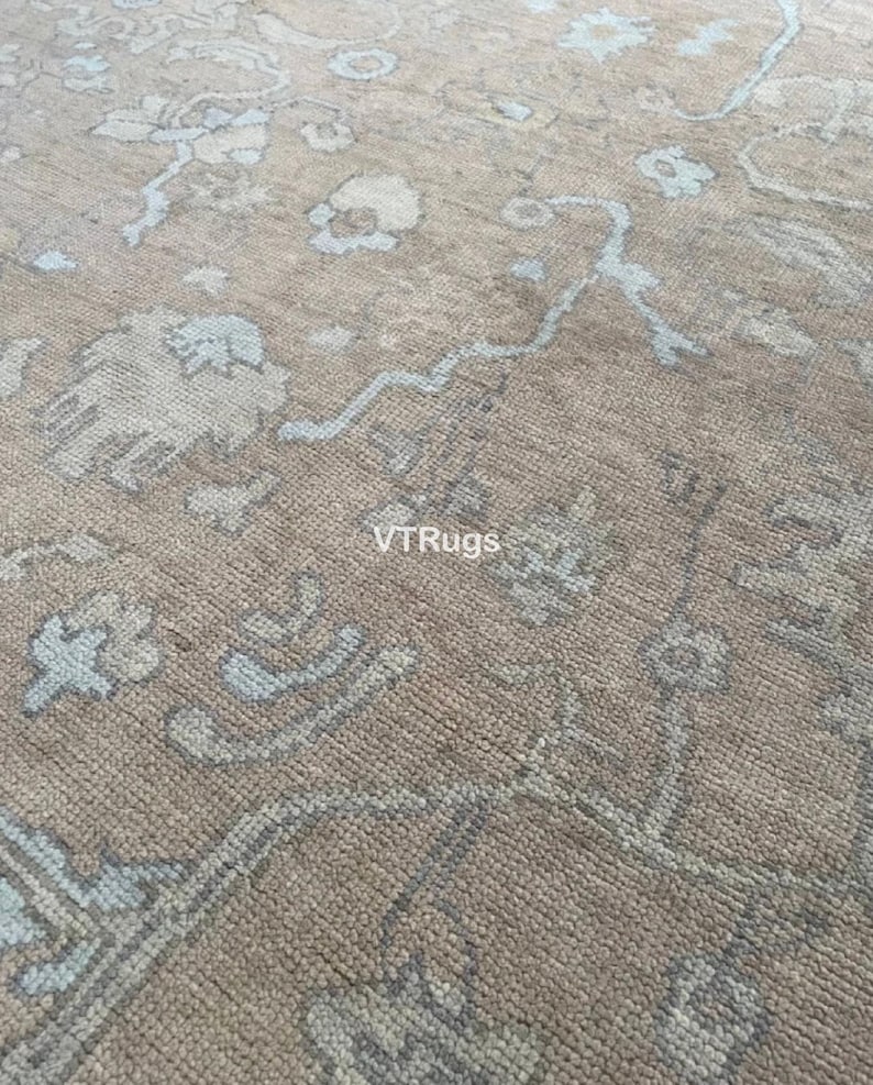 May include: Close-up of a beige rug with a detailed floral pattern in light blue and off-white. The rug has a textured appearance and the text "VTRugs" is visible.