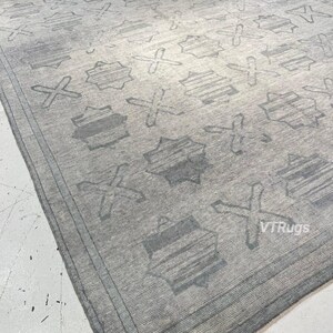May include: A large, rectangular gray rug with a repeating pattern of geometric shapes and crosses. The rug has a border and a textured appearance, suitable for a living room or bedroom.