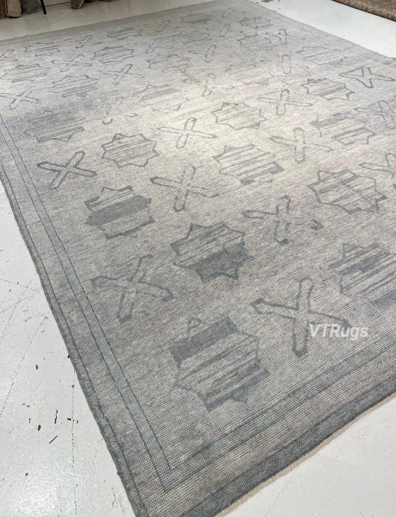 May include: A large, rectangular gray rug with a repeating pattern of geometric shapes and crosses. The rug has a border and a textured appearance, suitable for a living room or bedroom.