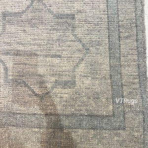 May include: Close-up of a light gray rug with a geometric pattern in shades of gray and blue. The rug has a textured surface and a border design. The text "VTRugs" is visible in the bottom right corner.