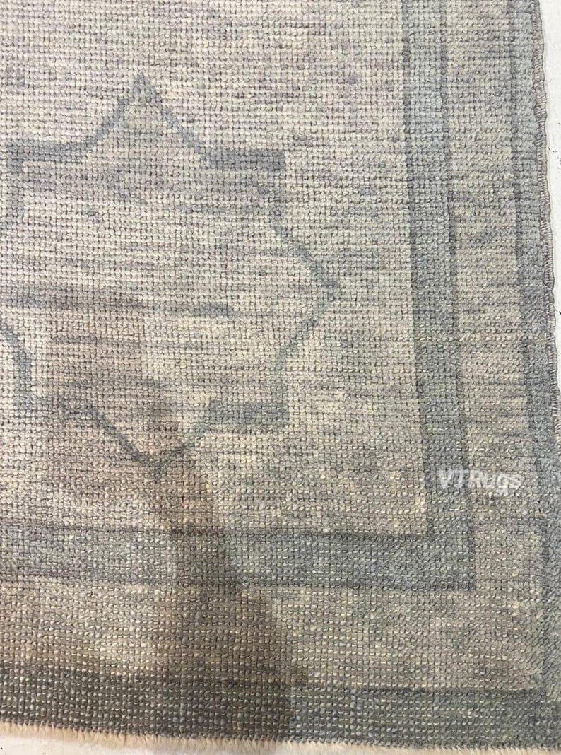 May include: Close-up of a light gray rug with a geometric pattern in shades of gray and blue. The rug has a textured surface and a border design. The text "VTRugs" is visible in the bottom right corner.