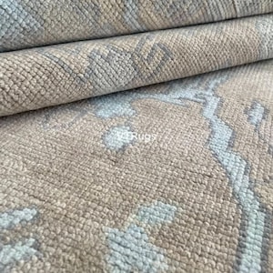May include: Close-up of a rolled-up rug with a textured surface. The rug features a beige background with a repeating pattern in light blue and gray. The rug's design is visible in the foreground, with the rolled layers in the background.