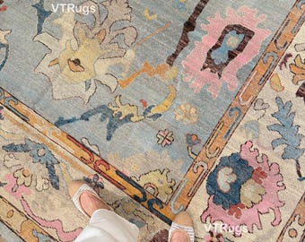 May include: A close-up shot of a light blue rug with a floral pattern in shades of pink, yellow, and brown. The rug has a border with a beige and orange design. The text "VTRugs" is visible on the rug.
