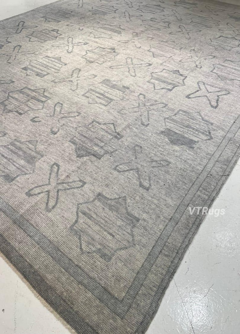 May include: A large, rectangular area rug in shades of gray. The rug features a repeating pattern of geometric shapes, including crosses and star-like figures. The border is a darker shade of gray, creating a defined edge.