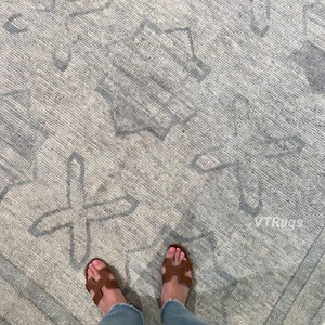 May include: A gray and white patterned rug with a geometric design. The rug has a textured surface and a border. Brown sandals and blue jeans are visible in the image.