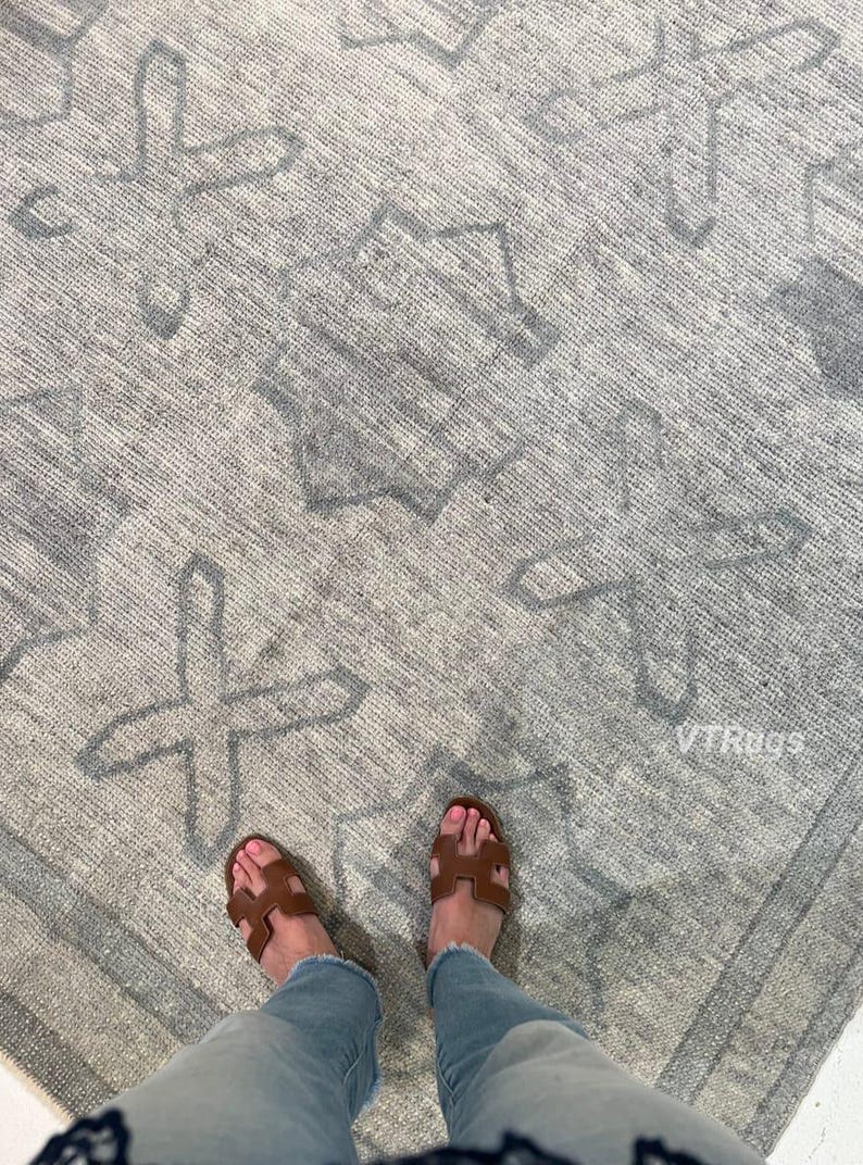 May include: A gray and white patterned rug with a geometric design. The rug has a textured surface and a border. Brown sandals and blue jeans are visible in the image.