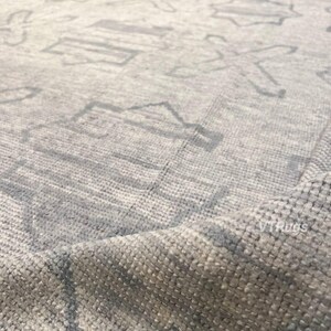 May include: Close-up of a light gray rug with a geometric pattern. The rug has a textured surface and a subtle color variation. The design features repeating shapes in a lighter shade of gray.