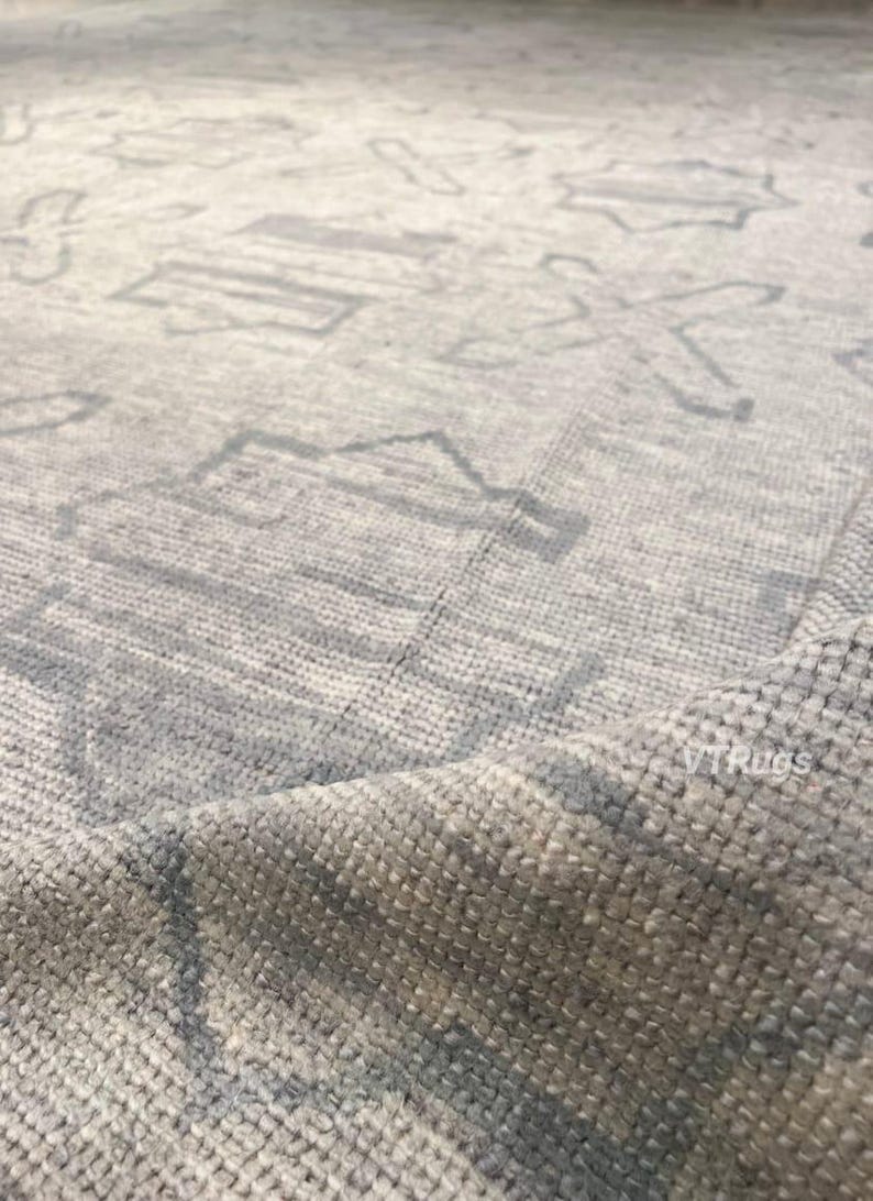May include: Close-up of a light gray rug with a geometric pattern. The rug has a textured surface and a subtle color variation. The design features repeating shapes in a lighter shade of gray.
