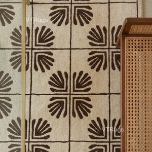 May include: Overhead shot of a beige rug featuring a repeating dark brown geometric design. Beside it, a gold-framed glass table and a wooden side table with a woven cane panel are visible.