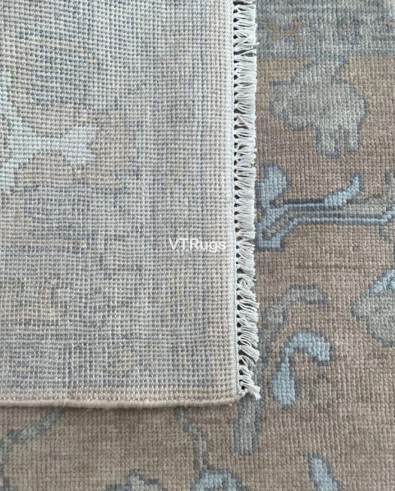 May include: Close-up of a beige and blue area rug with a floral pattern. The rug has a fringe edge and the text "VTRugs" is visible. The rug's design features light blue and beige floral motifs.