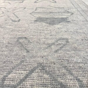 May include: Close-up of a light gray rug with a geometric pattern in a darker shade of gray. The rug's design features stylized shapes, including crosses and diamond-like figures, creating a textured surface.