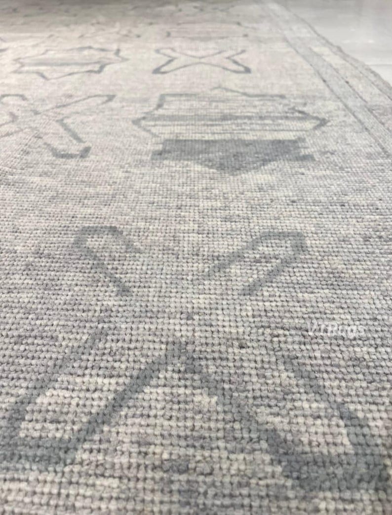 May include: Close-up of a light gray rug with a geometric pattern in a darker shade of gray. The rug's design features stylized shapes, including crosses and diamond-like figures, creating a textured surface.