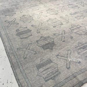May include: A large, rectangular area rug in shades of gray. The rug features a repeating pattern of geometric shapes, including star-like figures and cross-like designs. The rug has a textured appearance and a border.