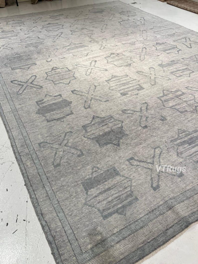 May include: A large, rectangular area rug in shades of gray. The rug features a repeating pattern of geometric shapes, including star-like figures and cross-like designs. The rug has a textured appearance and a border.