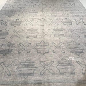 May include: A large, rectangular area rug in shades of gray. The rug features a repeating pattern of geometric shapes, including crosses and star-like designs. The overall design is symmetrical and the rug appears to be made of a woven material.