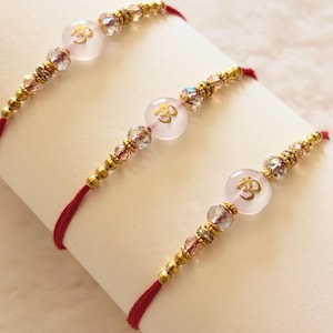 May include: Three red thread bracelets featuring a central pale pink circular bead with a gold Om symbol. Each bracelet is accented with small metallic gold beads and clear faceted crystal beads arranged symmetrically on either side of the centre.