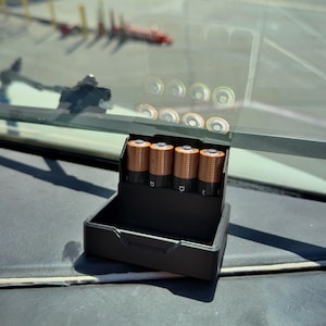 May include: A black plastic battery holder containing four copper-colored AA batteries. The batteries are arranged in a row, with the positive terminals visible. The holder sits on a dark surface, with a blurred outdoor scene visible through a window.