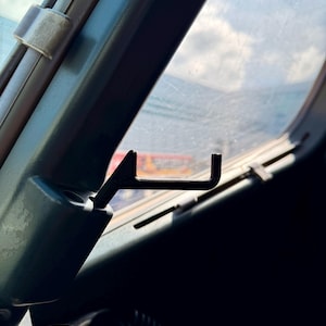 May include: Interior view of a vehicle's window frame, featuring a black metal hook and a window latch. The frame is a dark teal color, with a glimpse of the sky and clouds visible through the glass.