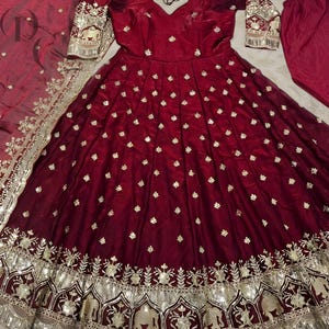 May include: A burgundy-colored formal dress with gold embroidery and embellishments. The dress features a V-neckline, long sleeves, and a full skirt. The skirt's hemline is adorned with intricate gold patterns and sequins. A matching scarf and pants are also included.