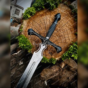 Può includere: Spada decorativa con impugnatura e guardia nere, con intricati disegni argentati. La lama presenta un motivo unico. La spada è appoggiata su una superficie di legno con fogliame verde.