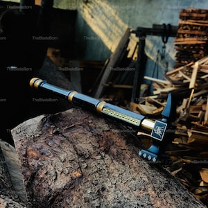 May include: A black and gold axe with a detailed design, resting on a weathered log. The axe features a long handle and a decorative head with intricate patterns. The blade is black, and the accents are gold.