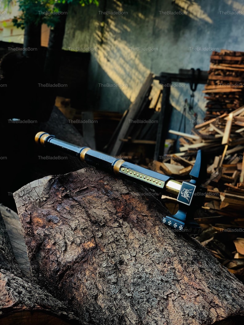 May include: A black and gold axe with a detailed design, resting on a weathered log. The axe features a long handle and a decorative head with intricate patterns. The blade is black, and the accents are gold.