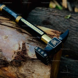 May include: A black and gold walking stick with a hammer-like head. The stick features gold accents and a decorative emblem on the head. The head has a cross design. The stick is resting on a wooden surface.
