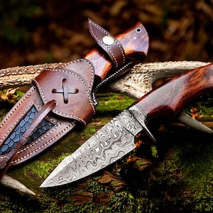 May include: A hunting knife with a patterned steel blade and a wooden handle. The knife is next to its brown leather sheath, which has a decorative cross-shaped design. The knife and sheath are displayed on a natural, mossy surface.