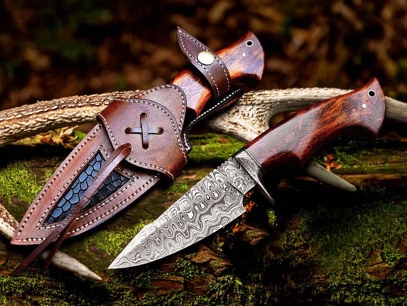 May include: A hunting knife with a patterned steel blade and a wooden handle. The knife is next to its brown leather sheath, which has a decorative cross-shaped design. The knife and sheath are displayed on a natural, mossy surface.