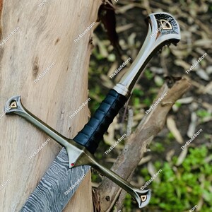 Könnte beinhalten: Detailliertes Schwert mit gemusterter Klinge und einem mit schwarzem Leder umwickelten Griff. Der Knauf ist silber- und goldfarben mit dekorativen Elementen. Der Text "TheBladeBorn" ist sichtbar. Das Schwert wird vor einem natürlichen Hintergrund präsentiert.
