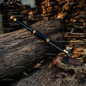May include: A black and gold battle hammer with an ornate design. The hammer is placed on a large, textured log, with a backdrop of stacked firewood. The hammer's head has a decorative pattern and the text "TheBladeBorn".