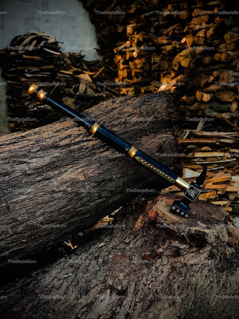 May include: A black and gold battle hammer with an ornate design. The hammer is placed on a large, textured log, with a backdrop of stacked firewood. The hammer's head has a decorative pattern and the text "TheBladeBorn".