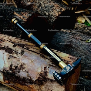 May include: A black and gold hammer with detailed embellishments, including an emblem and cross designs. The hammer's handle is black with gold accents. The hammer is placed on a wooden surface.