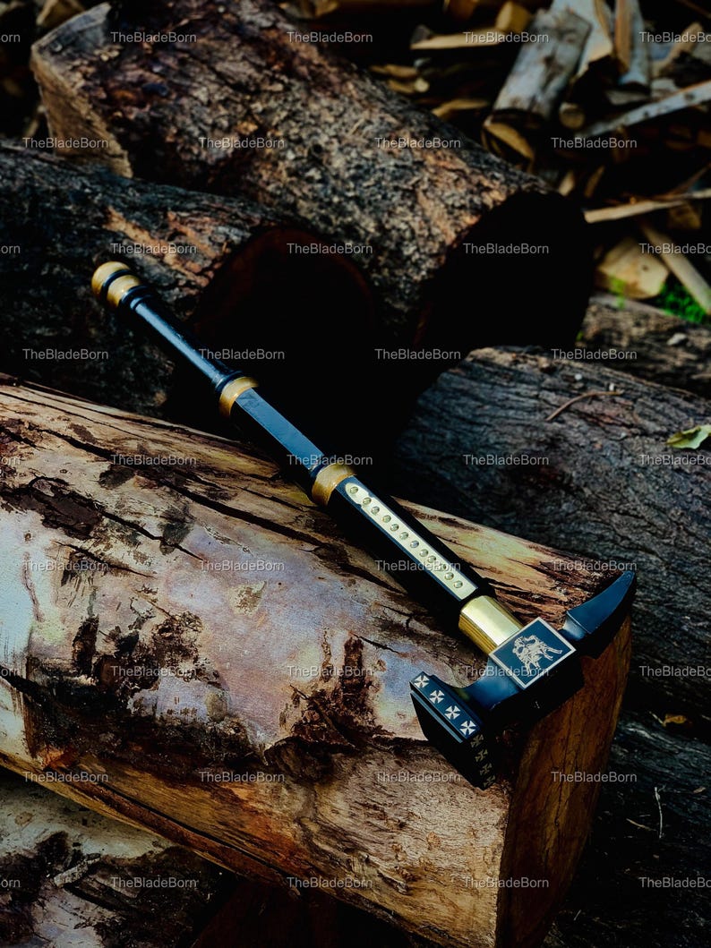 May include: A black and gold hammer with detailed embellishments, including an emblem and cross designs. The hammer's handle is black with gold accents. The hammer is placed on a wooden surface.