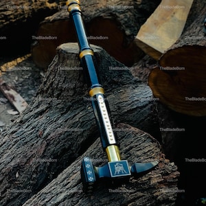 May include: A black and gold hammer with a long handle, resting on a pile of wood logs. The hammer head features a decorative emblem and cross symbols. The handle is black with gold accents.