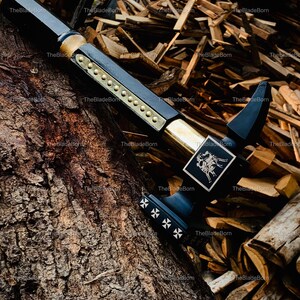 May include: A black and gold sword with intricate detailing. The sword has a black handle with gold embellishments and a decorative emblem. The blade is black with gold rivets and a cross design. The sword is displayed on a tree trunk.