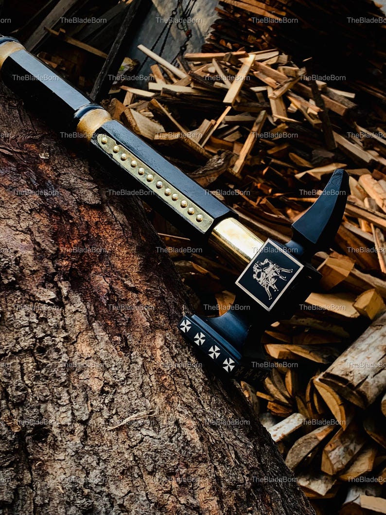 May include: A black and gold sword with intricate detailing. The sword has a black handle with gold embellishments and a decorative emblem. The blade is black with gold rivets and a cross design. The sword is displayed on a tree trunk.