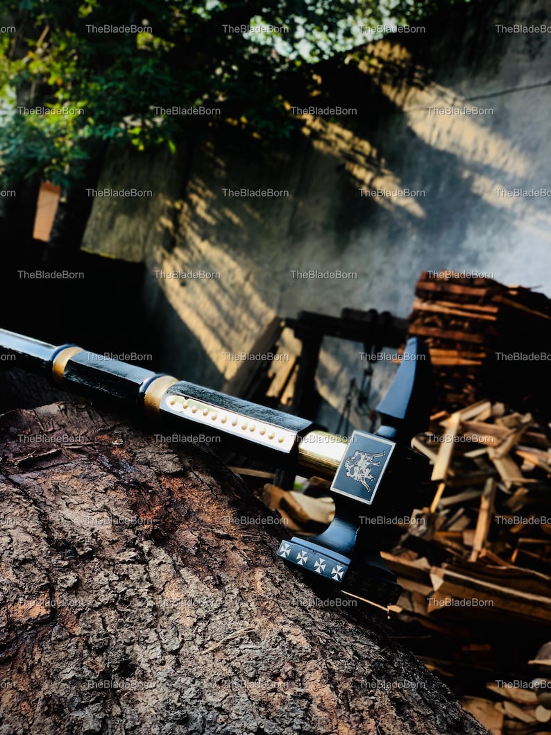 May include: A black and gold sword with a decorative handle, resting on a textured wooden surface. The sword features a detailed design on the handle and a silver blade. The background includes wood and foliage.