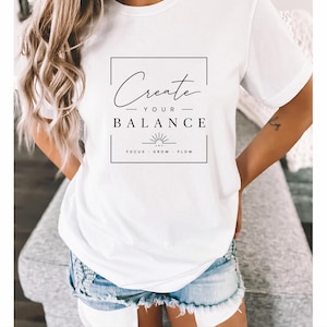 Könnte beinhalten: Weißes T-Shirt mit dem Text "Create Your Balance" in schwarzer Schrift in einem schwarzen rechteckigen Rahmen. Darunter stehen die Worte "Focus, Grow, Flow" und eine Sonnengrafik. Das Shirt ist aus weichem Material.