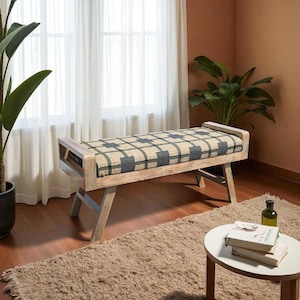 Peut inclure: Banc en bois avec coussin à carreaux. Le banc a un cadre en bois clair et des pieds inclinés. Le coussin présente un motif quadrillé beige, gris et noir. Le banc est placé devant une fenêtre avec des rideaux transparents. Le banc mesure environ 120 cm de long.